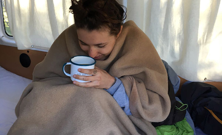 Een vrouw nestelt zich in een campervan in een deken en heeft een kopje thee in haar hand.